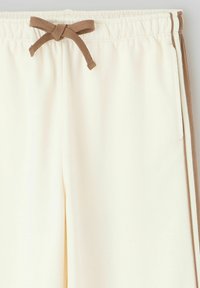 Joggers de couleur crème avec une taille élastique à cordon, ornés de rayures latérales marron et d'une texture de tissu douce et lisse.