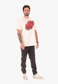 Uomo con barba e braccio tatuato che indossa una t-shirt bianca Diesel, jeans neri e sneakers bianche, in piedi contro uno sfondo bianco semplice.