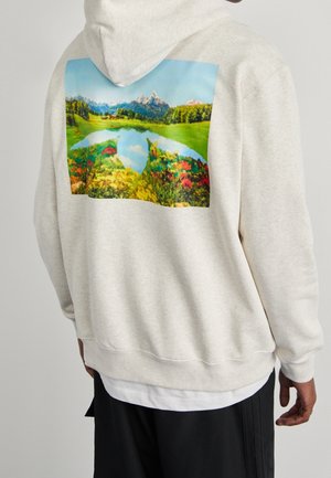 Osoba w beżowej bluzie z kapturem z kolorowym nadrukiem gór, jeziora i łąki na plecach, stojąca na jednolitym tle.