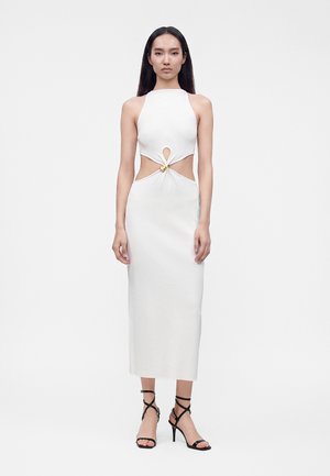Femme portant une robe midi blanche sans manches avec des découpes à la taille reliées par un anneau doré, associée à des talons hauts noirs à brides.