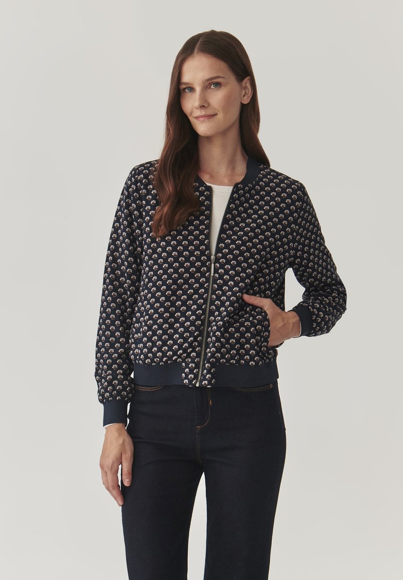 Chaqueta bomber azul con un patrón floral texturizado, puños y dobladillo acanalados, cierre frontal con cremallera y bolsillos laterales. Combinada con pantalones vaqueros oscuros.