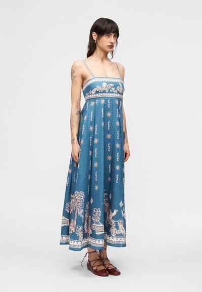 Γυναίκα που φοράει μπλε φόρεμα μέχρι τους αστραγάλους με λεπτές τιράντες και περίπλοκα λευκά και ροζ σχέδια, συνδυασμένο με σκούρα κόκκινα δετά παπούτσια.