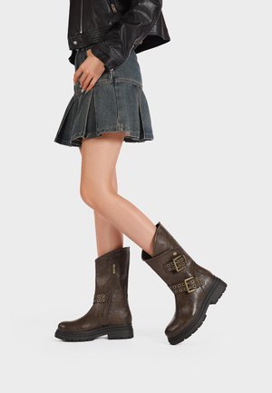 Stivaletti in pelle marrone con cerniere, due cinturini decorativi con fibbie, una finitura testurizzata e una suola nera spessa. Indossati con una gonna di jeans.