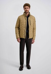 Gepolsterte Jacke in Khaki mit einem Zickzackmuster, mit Reißverschluss vorne und hohem Kragen. Kombiniert mit dunklen Hosen und Schuhen.