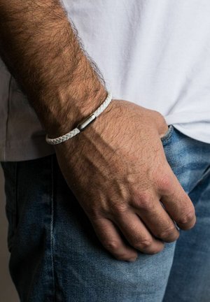 Pulsera de cuero trenzado blanco con un cierre cilíndrico metálico, llevada en una muñeca con vello visible, mano descansando sobre unos pantalones de mezclilla.