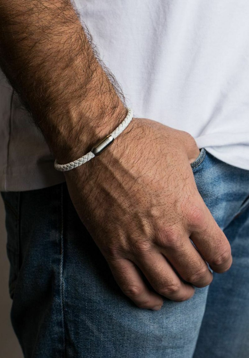 Bela pletena usnjena zapestnica z kovinskim cilindričnim sponko, nameščena na zapestju z vidnimi dlačicami, roka počiva na jeans hlačah.