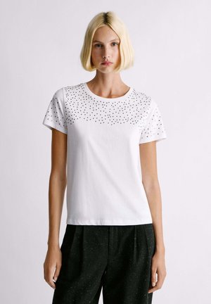 Mujer con cabello rubio corto, que lleva una camisa blanca de manga corta con pequeños puntos negros en el pecho y pantalones negros, de pie sin moverse.