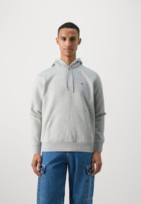 Sudadera gris de mezcla de algodón, con un bolsillo tipo canguro y capucha con cordón. Llevada con unos vaqueros cargo azules.