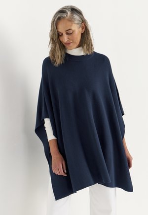 Vrouw met grijs haar die een marineblauwe poncho draagt over een witte coltrui en witte broek, kijkt naar beneden met een lichte glimlach.