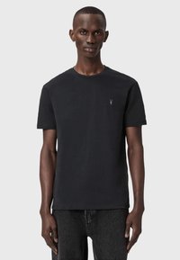 Musta crew neck t-paita, joka on valmistettu pehmeästä kankaasta ja jossa on hienovarainen logo rinnassa, yhdistettynä tummaan farkkuihin ja yksinkertaiseen taustaan.