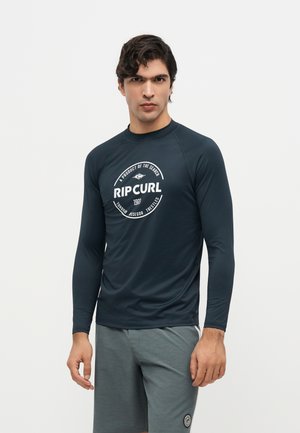Jeune homme portant un t-shirt Rip Curl à manches longues sombre et un short gris, debout devant un fond clair uni.