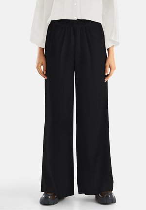 Black wide-leg trousers with a smooth, flowing fabric and an elastic waistband. Paired with a loose-fitting white shirt and chunky shoes.