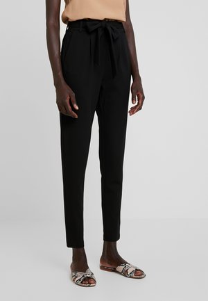 Osoba ubrana w czarne, zwężane spodnie z wysokim stanem i paskiem do wiązania oraz beżowe płaskie sandały z odkrytymi palcami w wężowy wzór na jasnym tle.