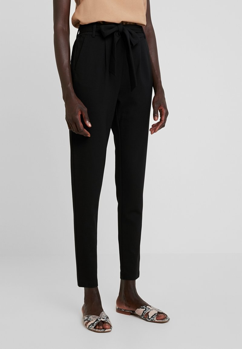 Personne portant un pantalon noir taille haute à jambes fuselées avec une ceinture nouée et des sandales plates à bout ouvert imprimé serpent beige sur un fond clair.