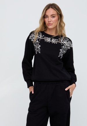 Frau trägt schwarzen Pullover mit weißer Blumenstickerei über den Schultern und schwarze Hose, Hände in den Taschen, neutraler Hintergrund.