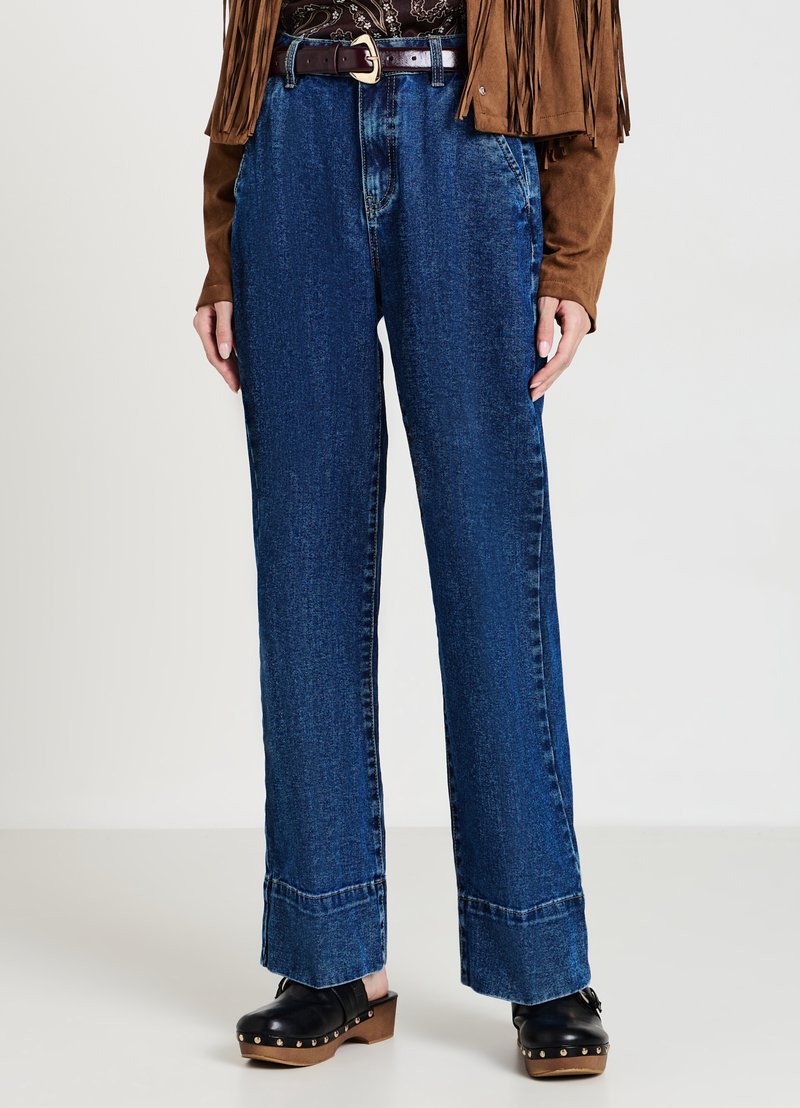 Jeans en denim bleu à taille haute et jambes larges, avec un ourlet cousu et une légère décoloration. Associés à des sabots noirs et une veste en daim marron.