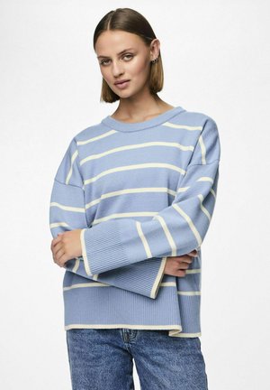Maglione a righe blu chiaro con righe gialle, vestibilità oversize, polsini e orlo a coste. Indossato con jeans in denim blu. Design rilassato.