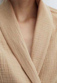 Beige teksturoitu kietaisutakki leveällä kauluksella, jossa on ryppyinen kangaspinta ja syvä V-aukko, joka luo pehmeän ja kevyen ilmeen.