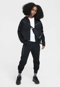 Tenue sportive noire comprenant un sweat à capuche zippé avec des accents gris et un pantalon de survêtement. Comprend un sac bandoulière noir et des baskets texturées.