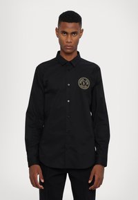 Camicia nera con bottoni, dotata di colletto, maniche lunghe e un logo rotondo dorato sul lato sinistro del petto. Il tessuto appare liscio e deciso.