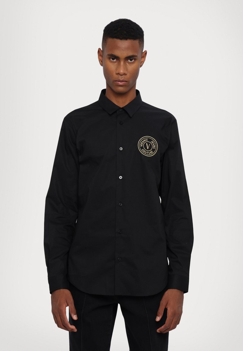 Camicia nera con bottoni, dotata di colletto, maniche lunghe e un logo rotondo dorato sul lato sinistro del petto. Il tessuto appare liscio e deciso.
