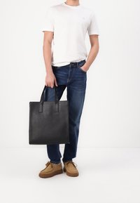 Person som har på sig en vit t-shirt, blå jeans och bruna skor, som håller en stor svart läder tote-väska i handtagen.