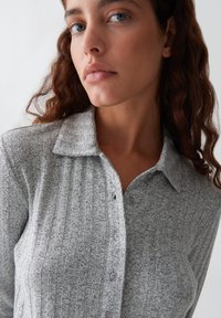 Camicia grigia con bottoni, realizzata in tessuto testurizzato con colletto e motivo a righe verticali. Presenta una leggera lucentezza e piccoli bottoni di colore chiaro.