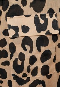 Tissu beige avec de grandes taches noires de léopard ; présente une poche poitrine avec un rabat. La texture semble lisse et légère.