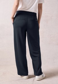 Tmavě modré, široké kalhoty s jemnou texturou, elastickým pasem a dvěma zadními kapsami, doplněné bílými teniskami.