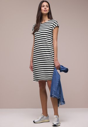 Joven mujer de pie sosteniendo una chaqueta de mezclilla azul, vistiendo un vestido de rayas blancas y negras hasta la rodilla y zapatillas plateadas contra un fondo liso.