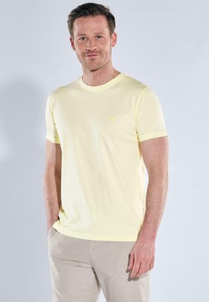 Mann trägt ein hellgelbes T-Shirt mit kurzen Ärmeln und beige Hose, steht mit einer Hand in der Tasche vor hellem Hintergrund.
