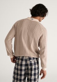 Beżowy sweter dziewiarski z detalami w warkocze, z prążkowanymi mankietami i kołnierzem. Połączony z kratkowymi spodniami w odcieniach granatu, kremu i szarości.