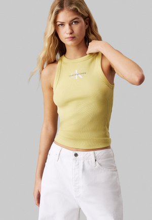 Giovane donna con lunghi capelli biondi indossa un top giallo smanicato Calvin Klein e pantaloni bianchi su uno sfondo chiaro e uniforme.