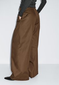 Hnědé široké kalhoty vyrobené z hladkého materiálu, s bočními kapsami a volným střihem. Doplňují je černobílé proužkované tričko.