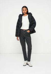 Čierna prešívaná bunda s prešívaným dizajnom, v kombinácii so šedými džínsami a bielymi členkovými čižmami. Model má na sebe biele tričko.