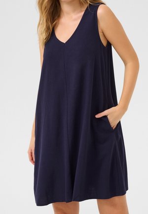 Femme portant une robe bleu marine sans manches, ample, avec un décolleté en V, la main droite placée dans la poche de la robe.