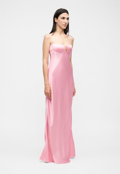 Γυναίκα στέκεται φορώντας ένα μακρύ ροζ σατέν φόρεμα με λεπτές τιράντες και δομημένη εφαρμογή στον μπούστο πάνω σε λευκό φόντο.