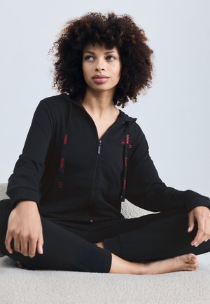 Jumpsuit noir zippé avec capuche, arborant des accents de logo rouges et des cordons de serrage ; texture douce ; modèle assis sur une surface claire.