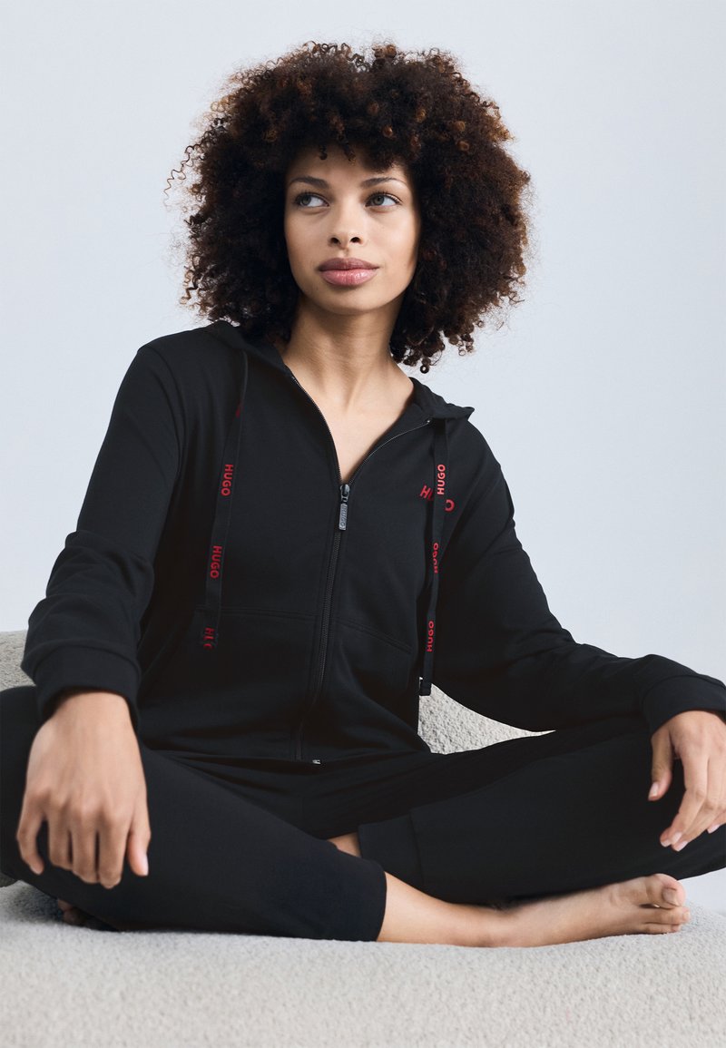 Svart zip-up jumpsuit med huva, med röda logotypdetaljer och dragsko; mjuk textur; modell sittande på en ljus yta.