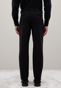 Uomo con pantaloni neri e scarpe nere, in piedi con le braccia rilassate ai lati contro un muro semplice e un pavimento rivestito in moquette.
