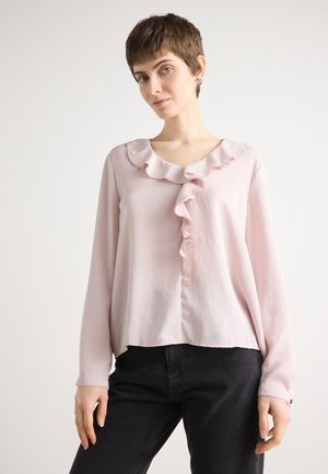 Lichtroze blouse met lange mouwen, gerimpelde halslijn en voorkant, zachte textuur, iets losvallende pasvorm, knoopdetail bij de manchetten, gecombineerd met zwarte jeans.