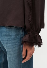 Main droite à côté d'un jean bleu portant un haut marron à manches longues avec un poignet plissé orné de dentelle et un détail de dentelle verticale sur la manche.