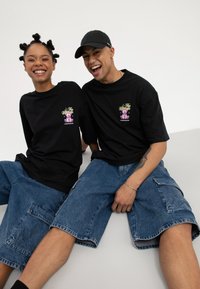 Două persoane zâmbitoare, așezate una lângă cealaltă, purtând tricouri negre cu imprimeu grafic și pantaloni scurți cargo din denim albastru, pe un fundal simplu.