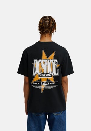 Osoba z krótkimi skręconymi włosami ma na sobie czarny T-shirt z dużą żółtą gwiazdą i napisem „DCSHOE COMPANY” na plecach, zestawiony z niebieskimi dżinsami.