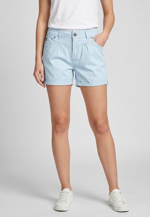 Woman wearing light blue cuffed shorts and white sneakers, standing with one hand in pocket against a plain background.