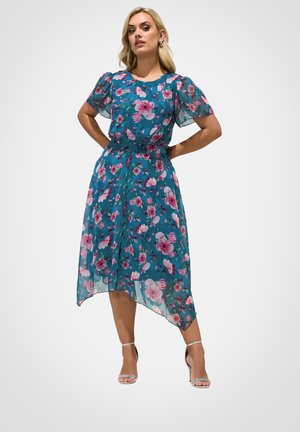 Kvinde står med hænderne på hofterne iklædt teal blomsterkjole med gennemsigtige korte ærmer og sølvfarvede ankelstrop hæle på neutral baggrund.