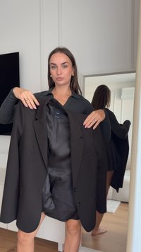 Zwarte blazer met satijnen binnenkant, revers met inkeping en twee knopen. Model houdt de blazer vast, draagt een geribd donker topje en staat voor een spiegelende achtergrond.