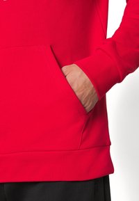 Červená mikina s kapucí z bavlněné směsi s klokaní kapsou. Má žebrované manžety a lem, s hladkou texturou a živou barvou.