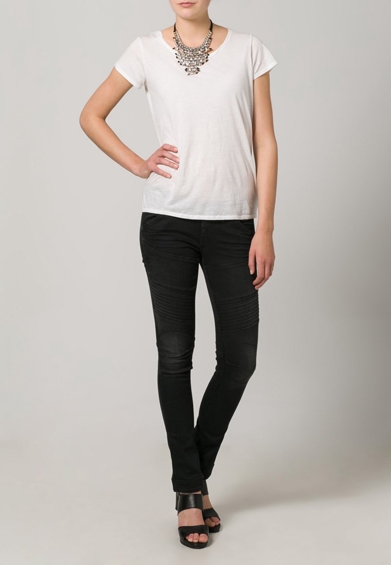 Camiseta blanca de manga corta combinada con jeans skinny negros con detalles acanalados, llevada con un collar plateado grueso y sandalias negras de tacón.