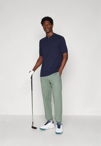 Jūras zilas krāsas polo kreklā ar īsām piedurknēm, kopā ar gaiši zaļām biksēm. Balti golfa apavi ar zilām detaļām. Tur rokā golfa nūju, stāv taisni.